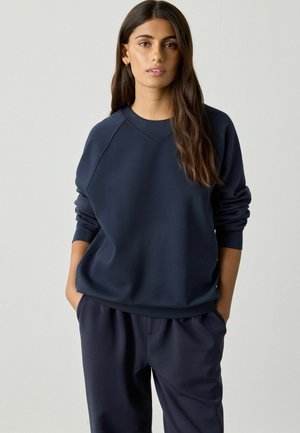 Woman with long brown hair wearing navy blue sweatshirt and matching pants, standing with hands in pockets against plain background.