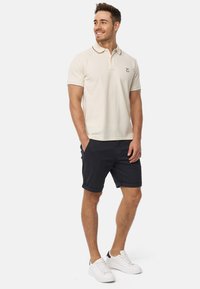 Beige Polohemd mit kontrastierendem Kragen und Ärmeln, kombiniert mit marineblauen Shorts. Das Modell trägt weiße Sneaker und steht mit den Händen in den Taschen.