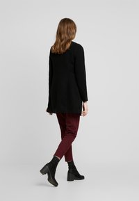 Vrouw die wegloopt in een zwarte jas, bordeauxrode broek en zwarte hoge enkellaarsjes tegen een effen grijze achtergrond.