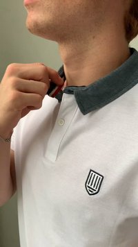 Bílá polo košile s kontrastním tmavě zeleným límcem, na hrudi s malým vyšitým logem ve tvaru štítu. Hladká textura, zapínání na knoflíky.