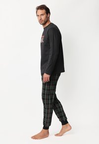 Hombre de pie descalzo, vistiendo una camiseta de manga larga negra y pantalones de pijama a rayas en negro, verde y rojo, frente a un fondo blanco.