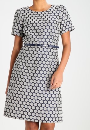 Frau trägt ein kurzärmeliges, knielanges Kleid mit weißem und dunkelblauem Kreis-Muster und einem schmalen dunkelblauen Gürtel in der Taille.