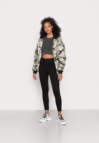 Kamuflāžas bomberis ar melniem akcentiem, pelēks īss tops, melnas šaurās bikses un masīvas kedas, stāvot ikdienišķā pozā uz neitrāla fona.