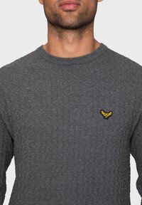 Suéter gris de punto con un acabado texturizado y un cuello redondo; presenta un pequeño logo bordado amarillo en el pecho.
