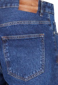 Jeans de mezclilla azul oscuro con un bolsillo trasero, que presentan costuras naranjas y una etiqueta de cuero marrón que dice "ZY PREMIUM."