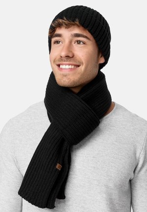Cappello in maglia nera a coste e sciarpa con finitura testurizzata. La sciarpa si adagia attorno al collo e presenta un piccolo dettaglio con etichetta marrone.