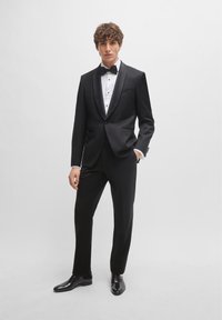 Black tuxedo with satin lapels, white dress shirt, and black bow tie. Fitted trousers and glossy black dress shoes complete the look.