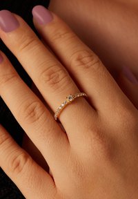 Gouden ring met een centrale steen, omringd door kleinere heldere stenen. Gladde band met een gepolijste afwerking die een hand met lavendelgekleurde nagels siert.