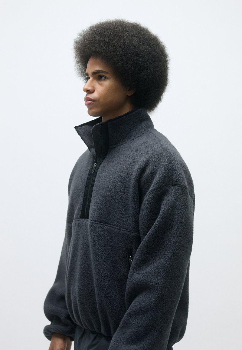 Fleece-pullover i mörkgrått med hög krage, halvdragkedja framtill och två sidofickor med dragkedja. Mjuk textur och avslappnad passform.