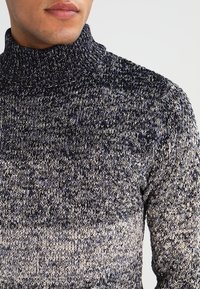 Gestrickter Rollkragenpullover in einer strukturierten Mischung aus dunklem Navy und Creme, mit engem Kragen und einem verlaufenden Muster nach unten.