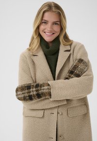 Beige uldfrakke med notcheskrålapel og to knapper, parret med en mørkegrøn turtleneck. Handsker med ternet mønster i brun, sort og creme.