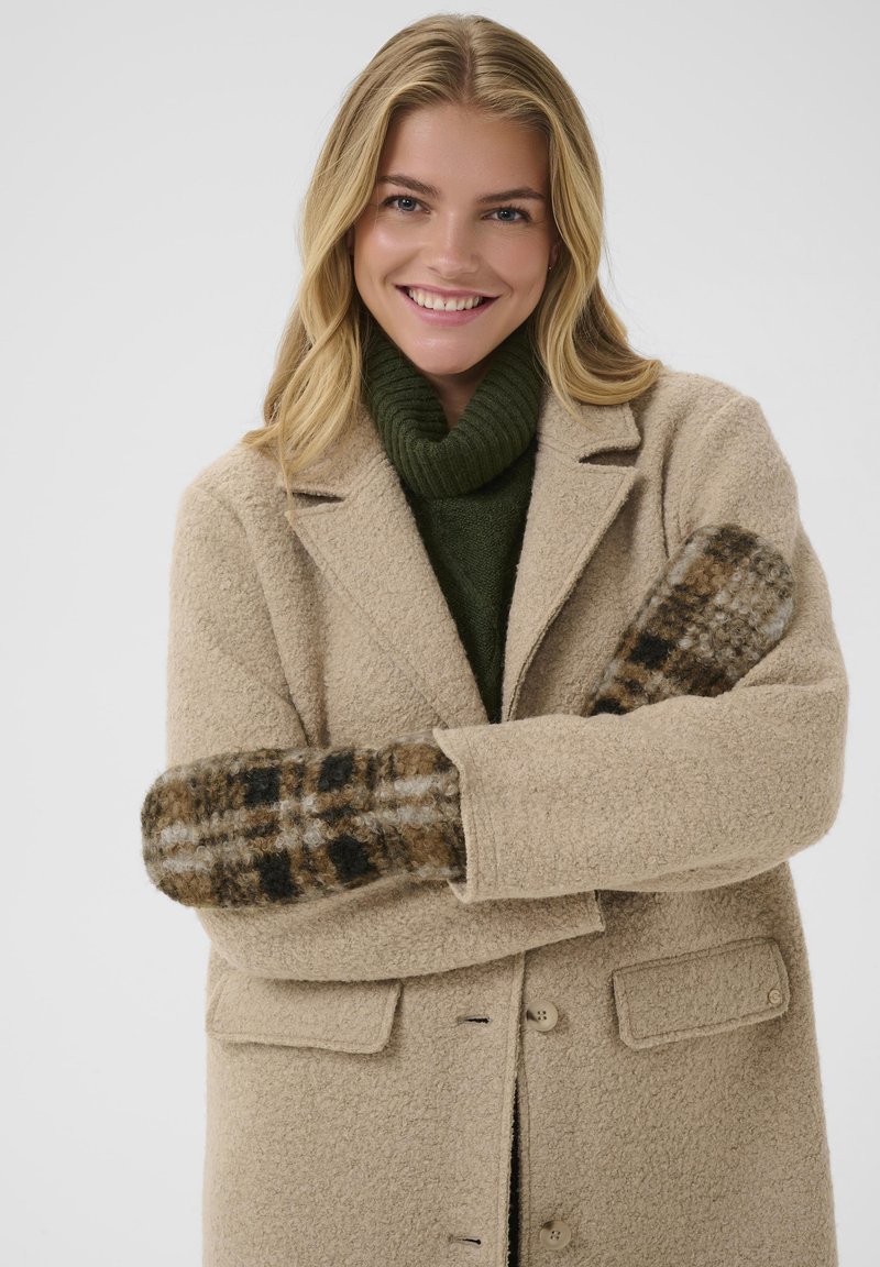Beige uldfrakke med notcheskrålapel og to knapper, parret med en mørkegrøn turtleneck. Handsker med ternet mønster i brun, sort og creme.