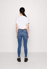 White T-shirt paired with blue skinny jeans. The jeans feature back pockets and a brown leather patch at the waistband. Black shoes complete the look.