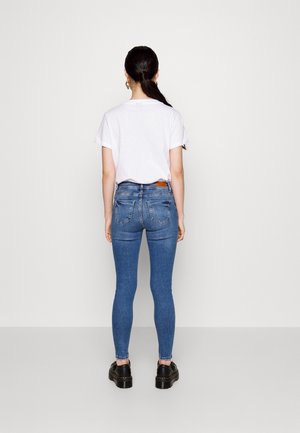 Camiseta blanca combinada con jeans ajustados azules. Los jeans tienen bolsillos traseros y un parche de cuero marrón en la cinturilla. Unos zapatos negros completan el look.