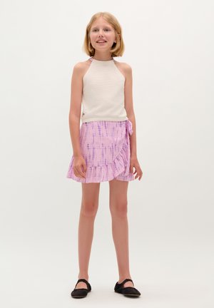 Witte gebreide mouwloze top gecombineerd met een roze tie-dye volantskirt. Zwarte platte schoenen maken de outfit compleet.
