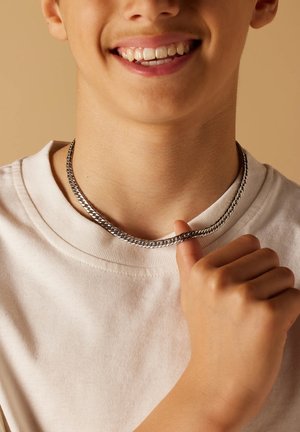 Zilveren ketting met een vlak, vergrendeld ontwerp, gedragen over een crèmekleurig shirt. De hand van het model houdt de ketting vast bij de hals.