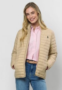 Bēša, quiltēta jaka ar izteiktu apkakli un smalkiem detaļām. Valkāta pāri gaiši rozā kreklam, apvienota ar ziliem džinsiem un bēšu jostu.