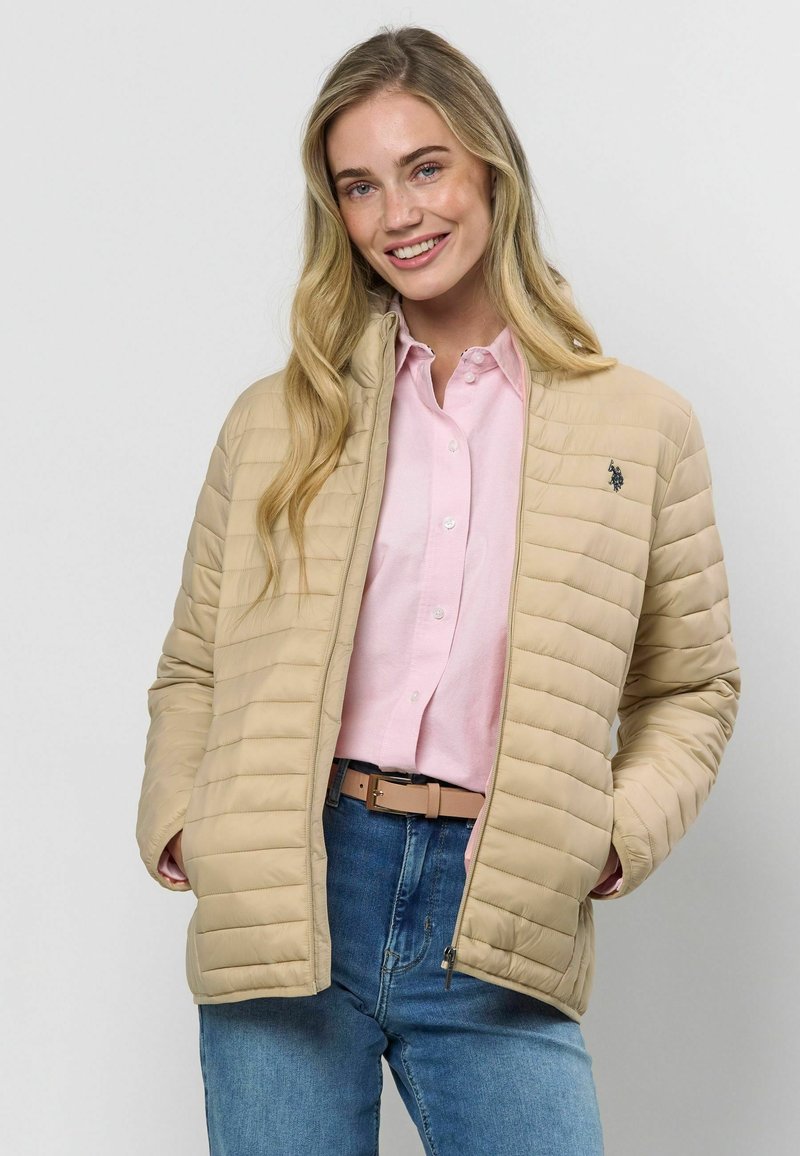 Bēša, quiltēta jaka ar izteiktu apkakli un smalkiem detaļām. Valkāta pāri gaiši rozā kreklam, apvienota ar ziliem džinsiem un bēšu jostu.