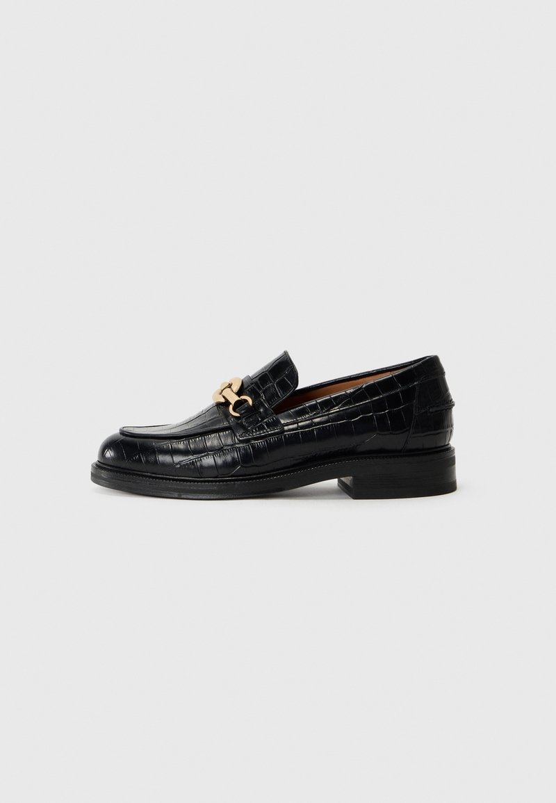Mocassin en cuir noir avec motif crocodile et décoration chaîne dorée sur semelle à talon bas sur fond gris clair.