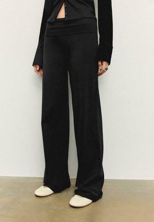 Osoba oblečená v čiernych širokých nohaviciach, čiernom dlhom rukáve s gombíkmi a bielych plochých topánkach stojaca na svetlej podlahe.