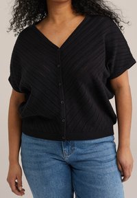 Žena v černém krátkém rukávu button-up kardiganu s texturovaným vzorem chevron a modrými džíny, stojící na jednobarevném pozadí.