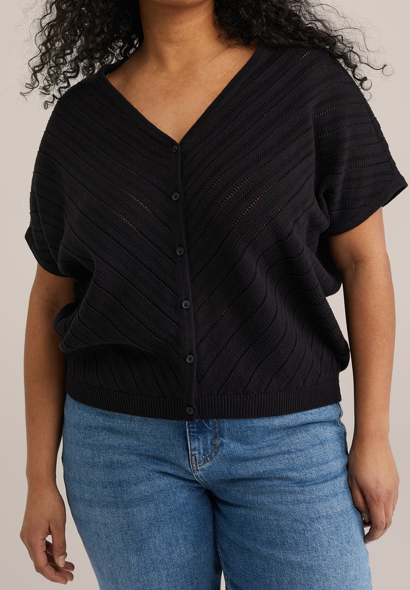 Žena v černém krátkém rukávu button-up kardiganu s texturovaným vzorem chevron a modrými džíny, stojící na jednobarevném pozadí.