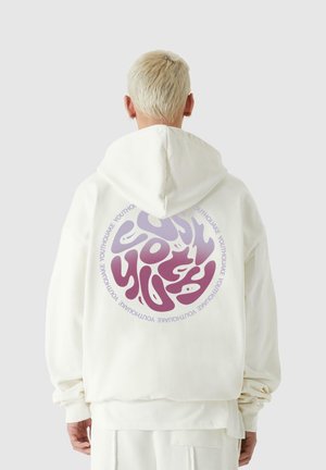 Osoba s krátkými blond vlasy nosící bílou mikinu s fialovým a růžovým psychedelickým kruhovým nápisem "YOUTHQUAKE" na zádech.
