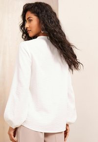 Blusa bianca a trama con maniche a palloncino, finitura liscia e scollatura rotonda, abbinata a pantaloni beige, vista da dietro.