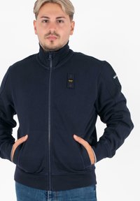 Marineblauwe ritsjack met hoge kraag, zijzakken en een textuurstof. Heeft een klein logo nabij de rits en ribgebreide manchetten.
