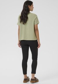 Top verde chiaro a maniche corte con una finitura testurizzata, abbinato a pantaloni neri slim-fit e scarpe marroni con allacciatura, visto da dietro.