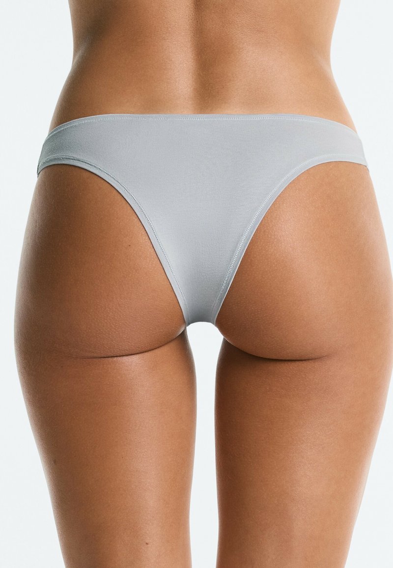 Vue arrière d'une personne portant un sous-vêtement bikini gris clair sans couture, montrant le bas du dos, les hanches et le haut des cuisses sur un fond uni.