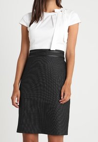 Haut blanc avec un détail nœud, associé à une jupe noire cintrée. Tissu lisse, manches courtes et une coupe jusqu'au genou avec une finition texturée.