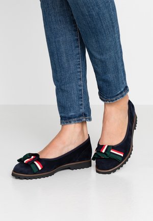 Granatowe baletki z zieloną i czerwoną wstążką w paski, noszone z dopasowanymi dżinsami. Tło jest jasne, w neutralnym kolorze.