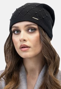 Zwarte beanie met een textuurpatroon en decoratieve zwarte kralen, afgewerkt met een klein metalen label met merknaam. Zacht gebreid materiaal.