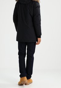 Svart isolerad jacka med en huva klädd i päls, kombinerad med mörka jeans och bruna stövlar. Synliga sömmar och detaljer i texturen.