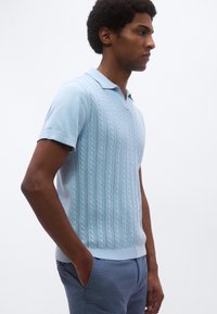 Ανοιχτό μπλε πλεκτό πόλο πουκάμισο με υφασματικό μοτίβο, κοντά μανίκια και γιακά που καμπυλώνει προς τα κάτω. Εφαρμόζει στενά στο σώμα.