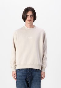 Kirkas beige huppari, jossa on pyöreä kaula-aukko ja pitkät hihat. Rinnassa on brodeerattu teksti, ja hihansuissa sekä helmassa on resorit. Yhdistetty sinisiin farkkuihin.