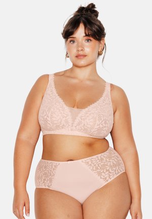 Donna con capelli scuri raccolti in uno chignon, che indossa un reggiseno in pizzo rosa chiaro abbinato e slip a vita alta, in piedi davanti a uno sfondo chiaro e uniforme.