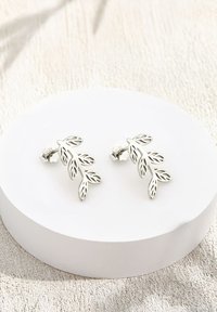 Silverblad-formade örhängen i stud-stil med intrikata utskärningar, med en texturerad yta och säker fästanordning, placerade på en rund utställning.