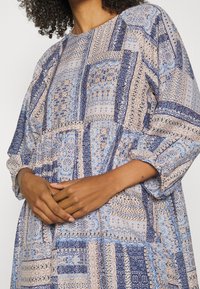Langärmliges Kleid mit einem Patchwork-Design in Blau, Beige und Creme, aus leichtem Stoff mit aufwendigen Mustern gefertigt.
