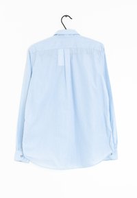 Camisa de manga larga de color azul claro con cuello abotonado, dobladillo curvado y puños abotonados, hecha de una tela texturizada. Cuelga de un gancho negro.