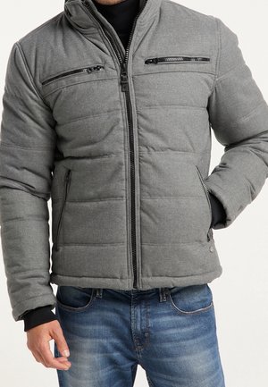 Homme portant une veste matelassée grise avec des poches à fermeture éclair noires, des poignets intérieurs noirs, un jean bleu, main dans la poche de la veste.