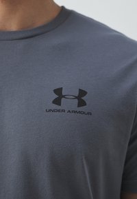 Camiseta de algodón gris con mangas cortas que presenta un logo negro de Under Armour en el pecho. Textura suave y corte estándar.