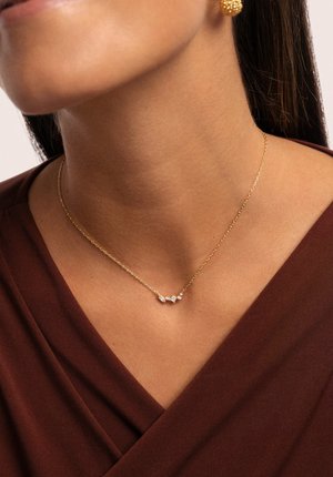 Collier en or avec trois charms géométriques ornés de pierres transparentes, reposant sur un haut marron. Texture lisse, design délicat.