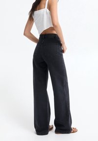 Jeans en denim noir à taille haute et jambes larges, avec deux poches arrière et des détails de couture visibles. Portés avec un top court blanc et des sandales.