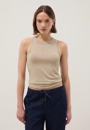 Mujer joven con cabello corto castaño, vestida con top sin mangas beige y pantalones azul oscuro con cordón, de pie contra un fondo claro y liso.