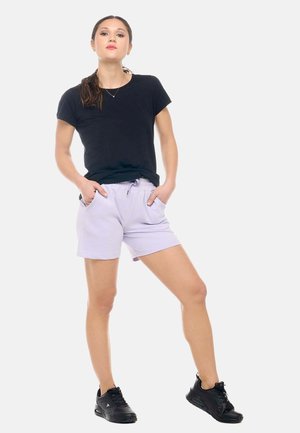 Femme debout les mains dans les poches, portant un t-shirt noir, un short violet clair et des baskets noires sur fond blanc.