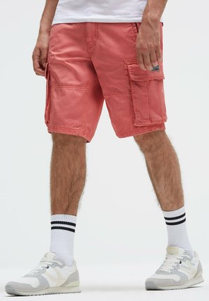 Mann trägt korallfarbene Cargo-Shorts, weiße Turnschuhe und weiße Socken mit zwei schwarzen Streifen, steht vor einem einfarbigen Hintergrund.