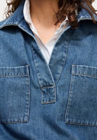 Denimshirt met een diepe kraag, twee voorzakken en contrasterende stiksels. De stof heeft een textuur en is medium blauw van kleur.