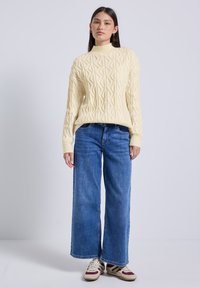 Jeune femme debout, portant un pull en laine écru à mailles torsadées, un jean bleu à jambes larges et des baskets blanches avec des rayures rouges et vertes.